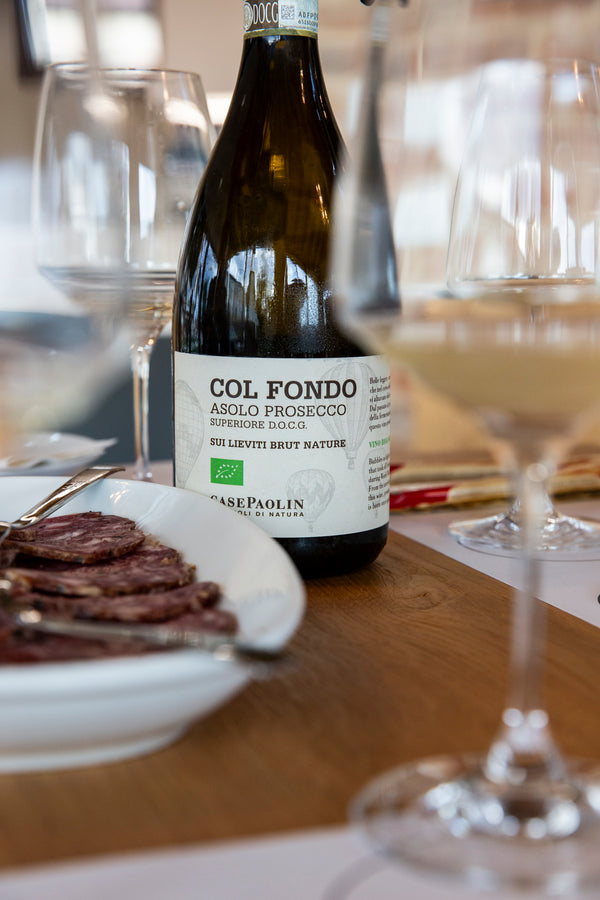 Bild einer Flasche Col Fondo Asolo Prosecco hinter einer Platte Salami zwischen Weingläsern durchfotografiert.