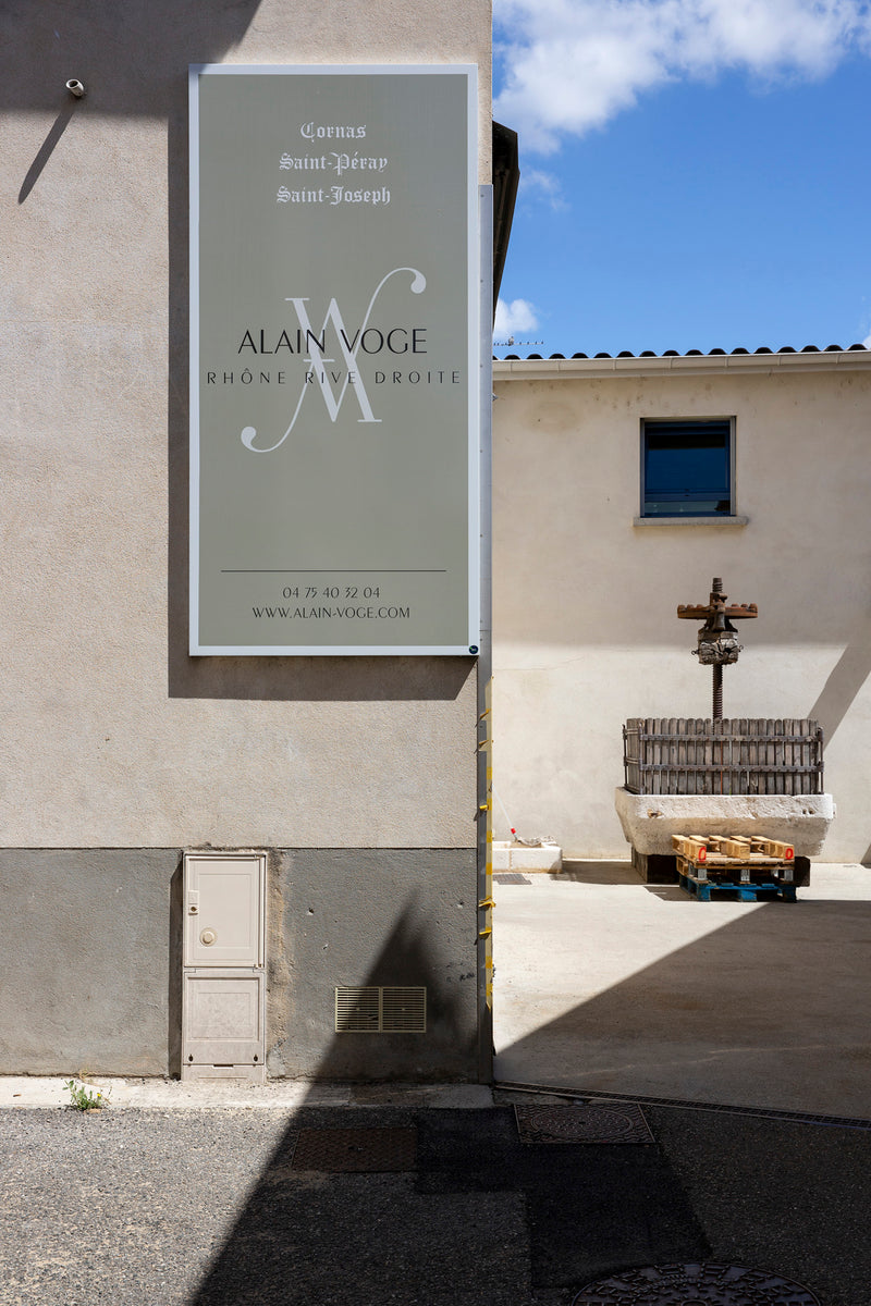Das Firmenschild der Domaine Alain Voge an einer Hauswand. Im Hintergrund ist eine alte eckige Korbpresse zu sehen.