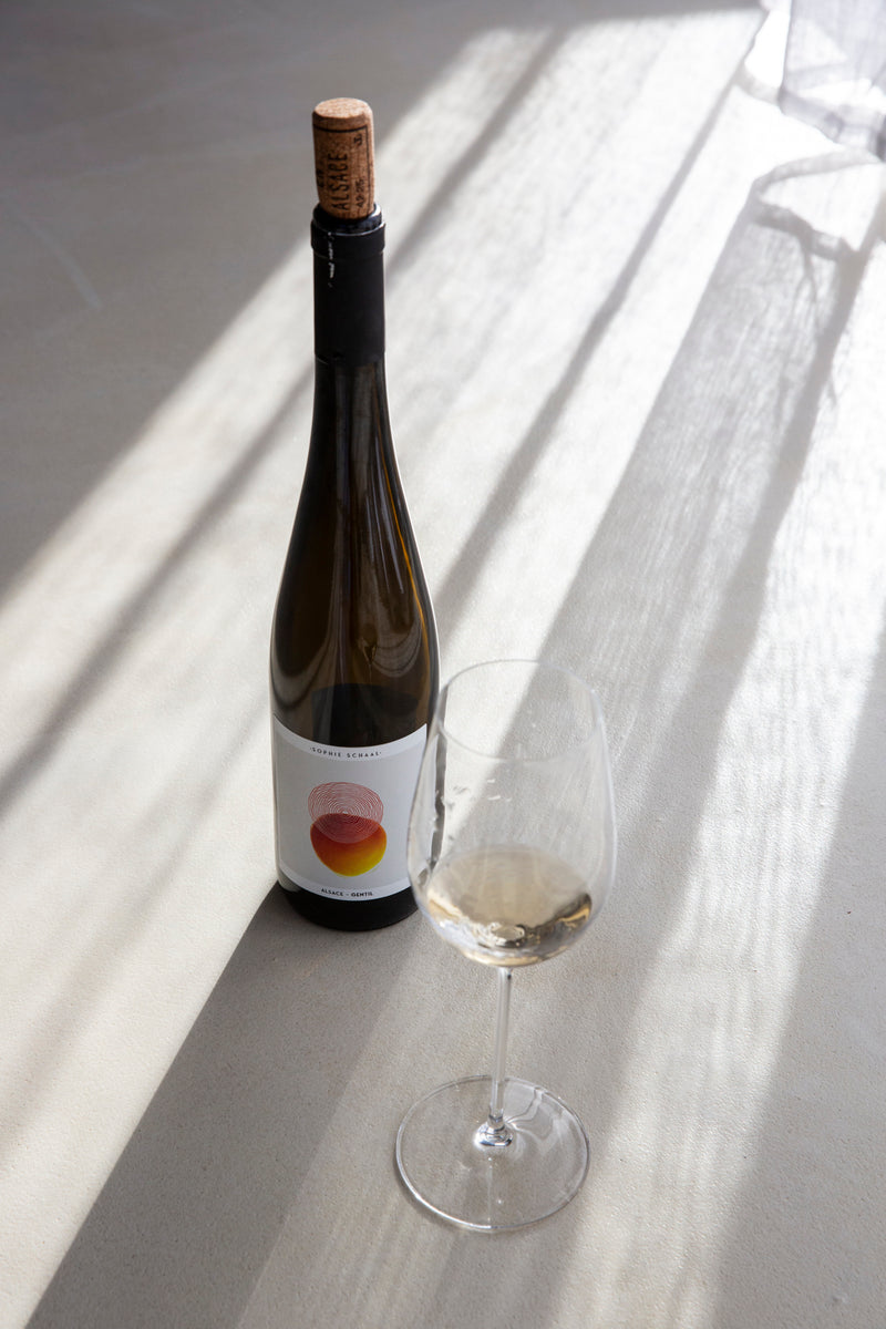 Foto einer Flasche Wein von Sophie Schaal, mit einem befüllten Weissweinglas, im zarten Schattenspiel eines hellen Vorhangs.